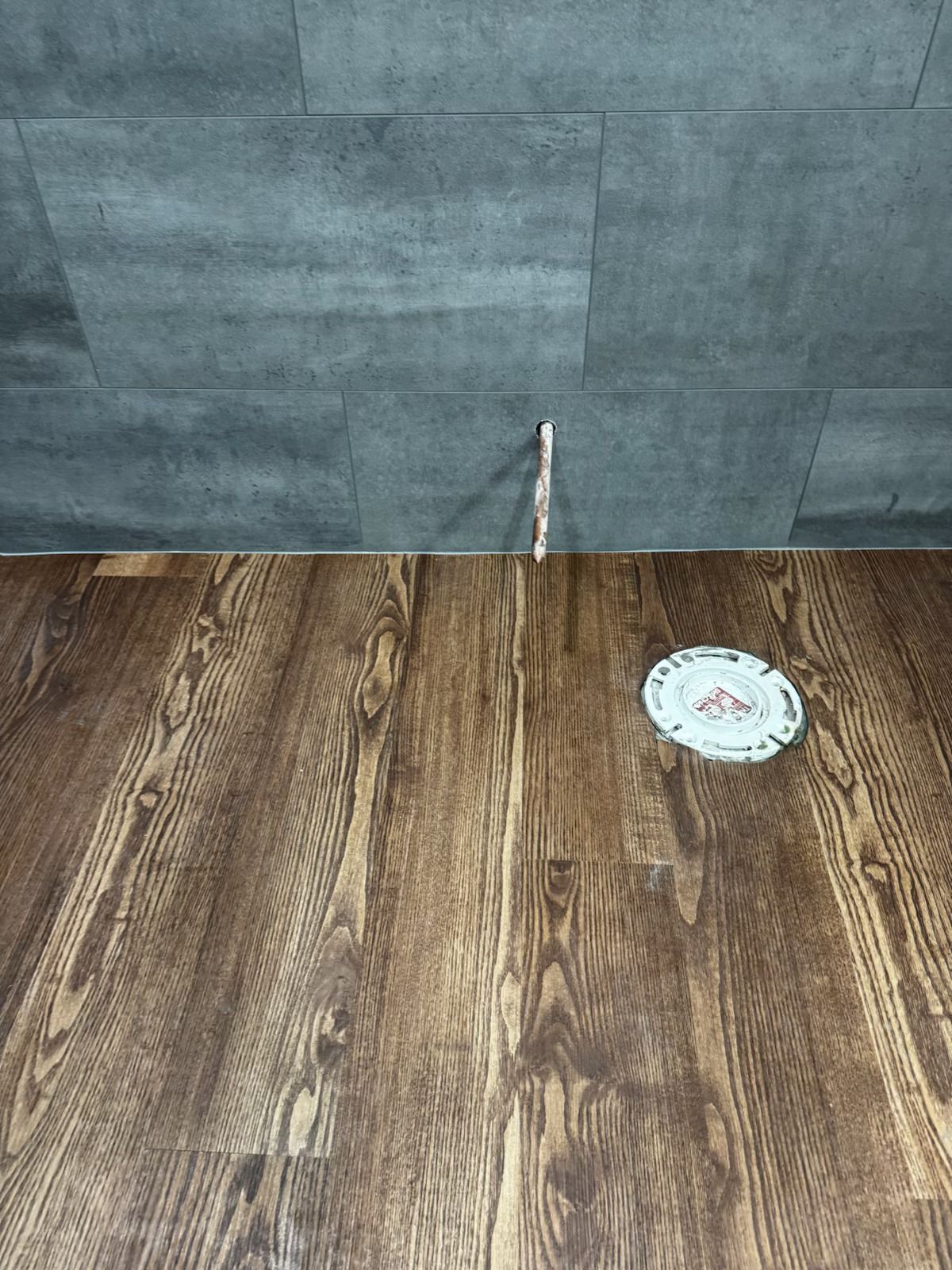 Wood-look floor meeting gray tile wall, with a small round wall fixture near the bottom right.