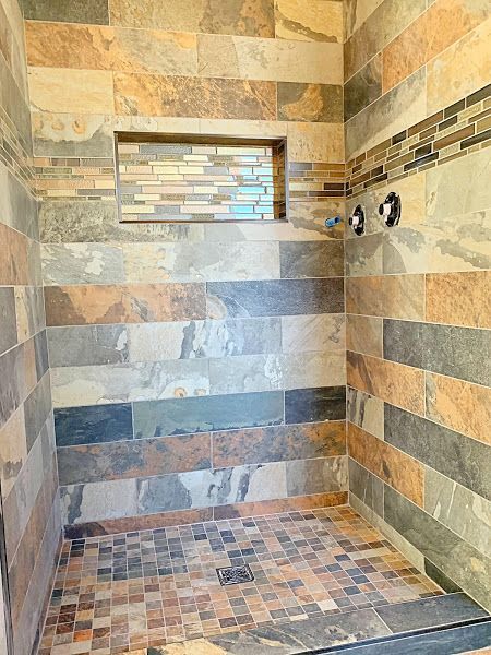 Tiled walk-in shower with mosaic floor, striped stone walls, recessed shelf, and dual shower controls.