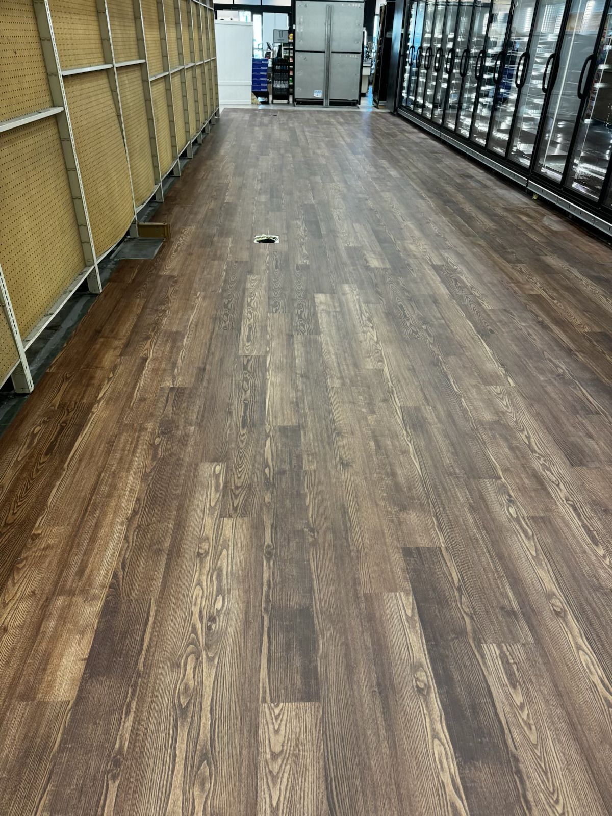 A grocery store aisle with wood-look flooring, empty shelves on the left, and refrigerated cases on the right.