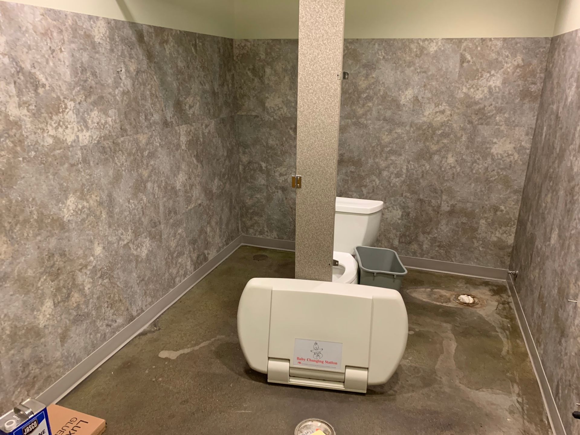 Empty restroom stall with gray textured walls, toilet, and a mounted baby changing station in the foreground