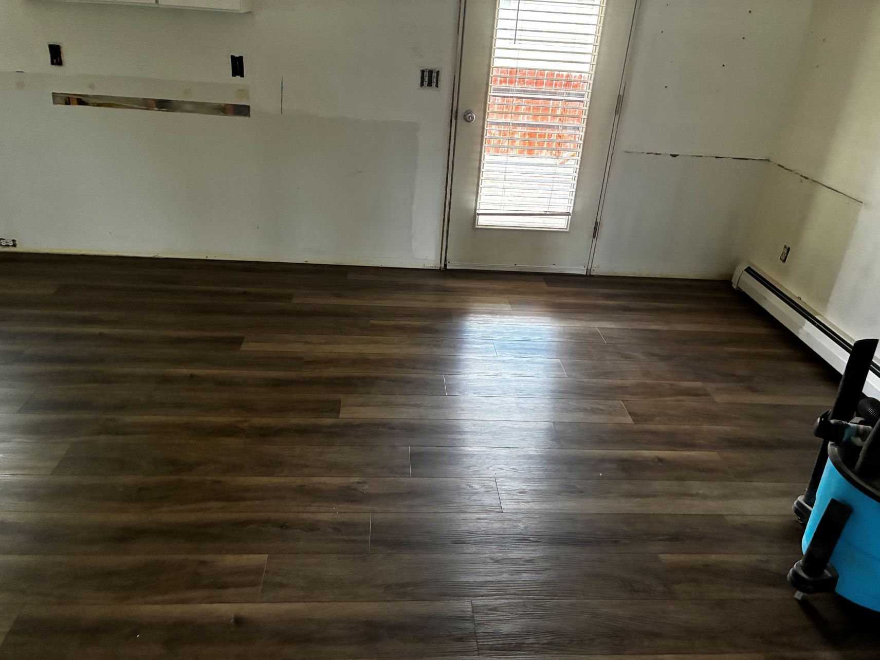 Empty room with dark wood flooring, white walls, a bright window, and a vacuum on the right.