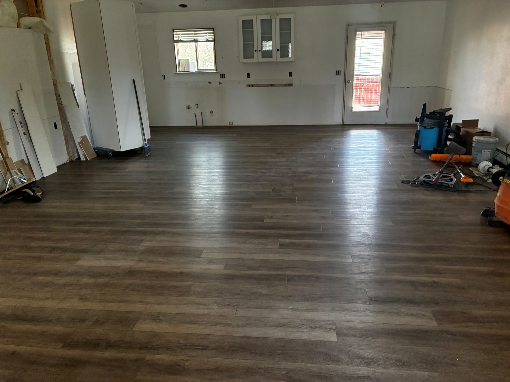 Empty room with wood-look flooring, white walls, windows, and tools stacked along the right side.