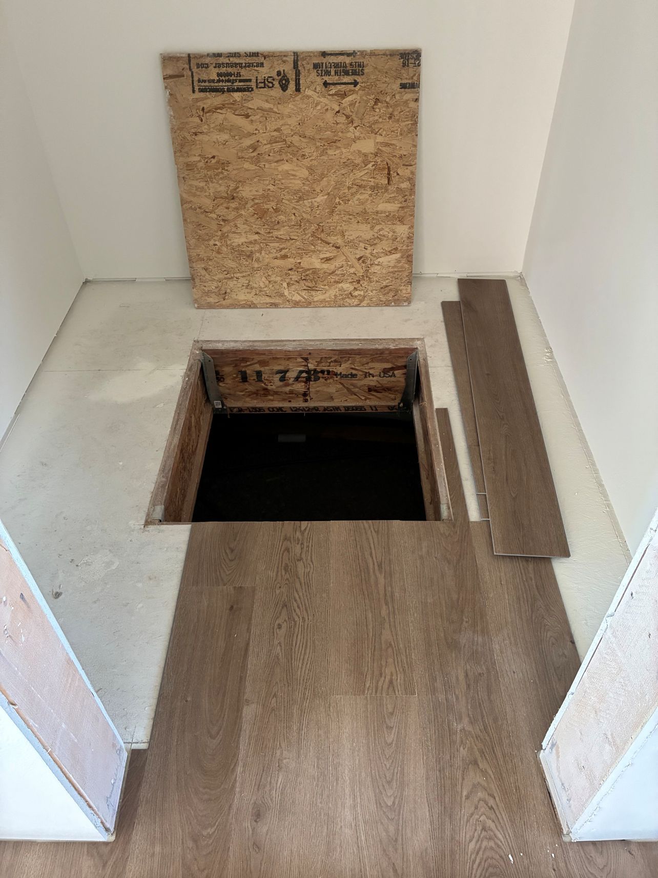 Open floor hatch in a small room with plywood cover and dark opening in the wooden floor