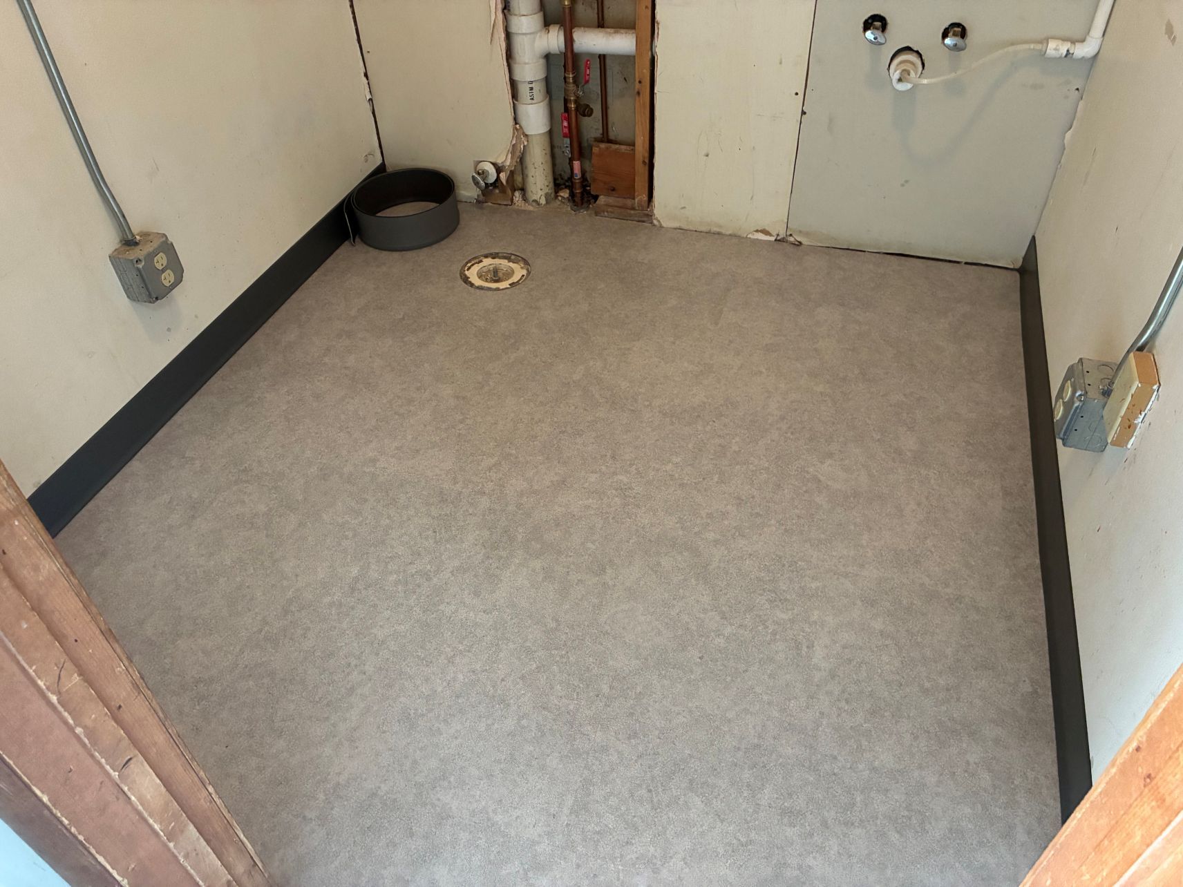 Small empty utility room with gray carpet, white walls, pipes, and a floor drain.