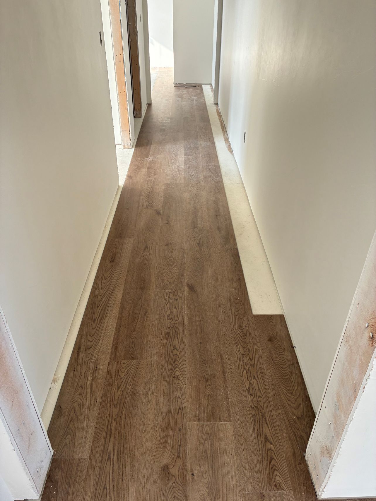 Narrow hallway with white walls and wood flooring, leading to a bright doorway.
