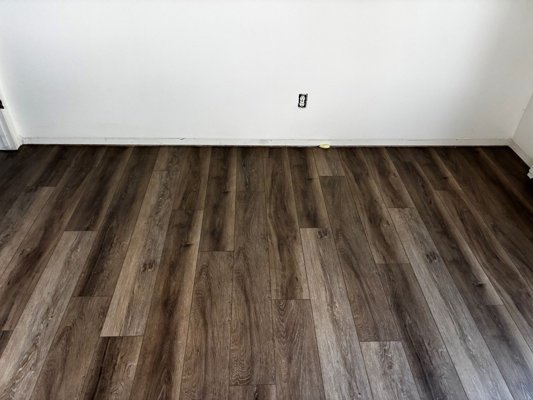 Empty room with white walls and dark wood flooring, showing a wall outlet.