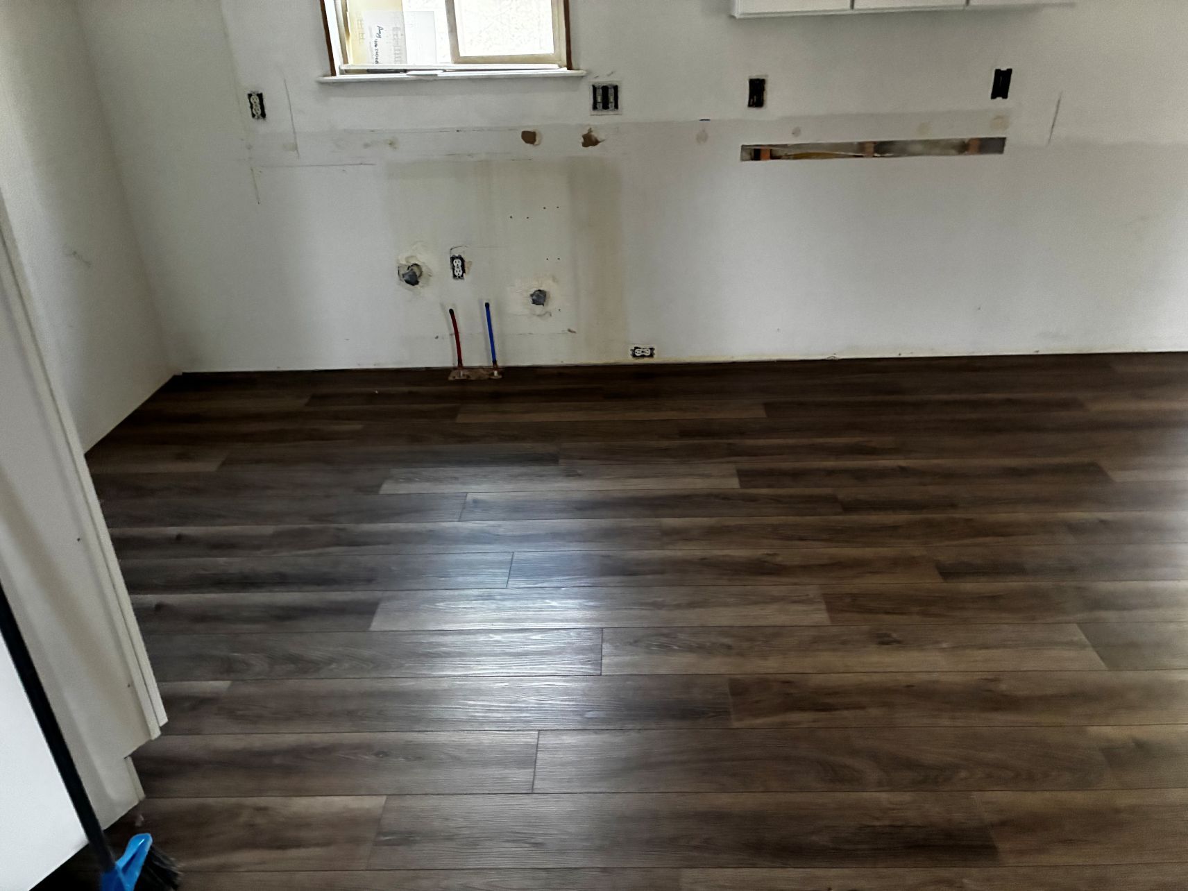 Empty room with dark wood flooring, white walls, and kitchen hookups under a window.