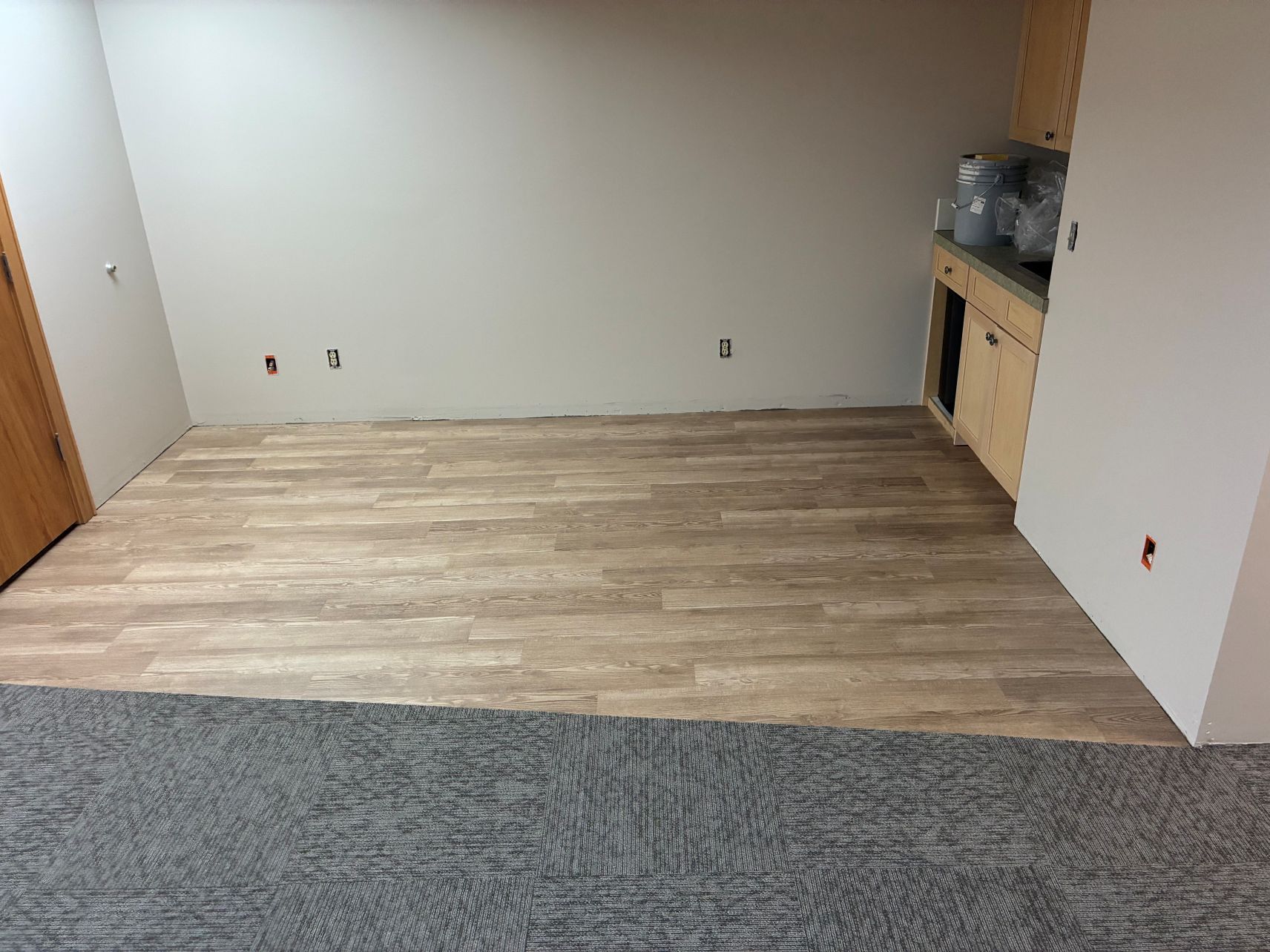 Empty room with wood-look flooring, gray carpet edge, and a small kitchenette on the right.