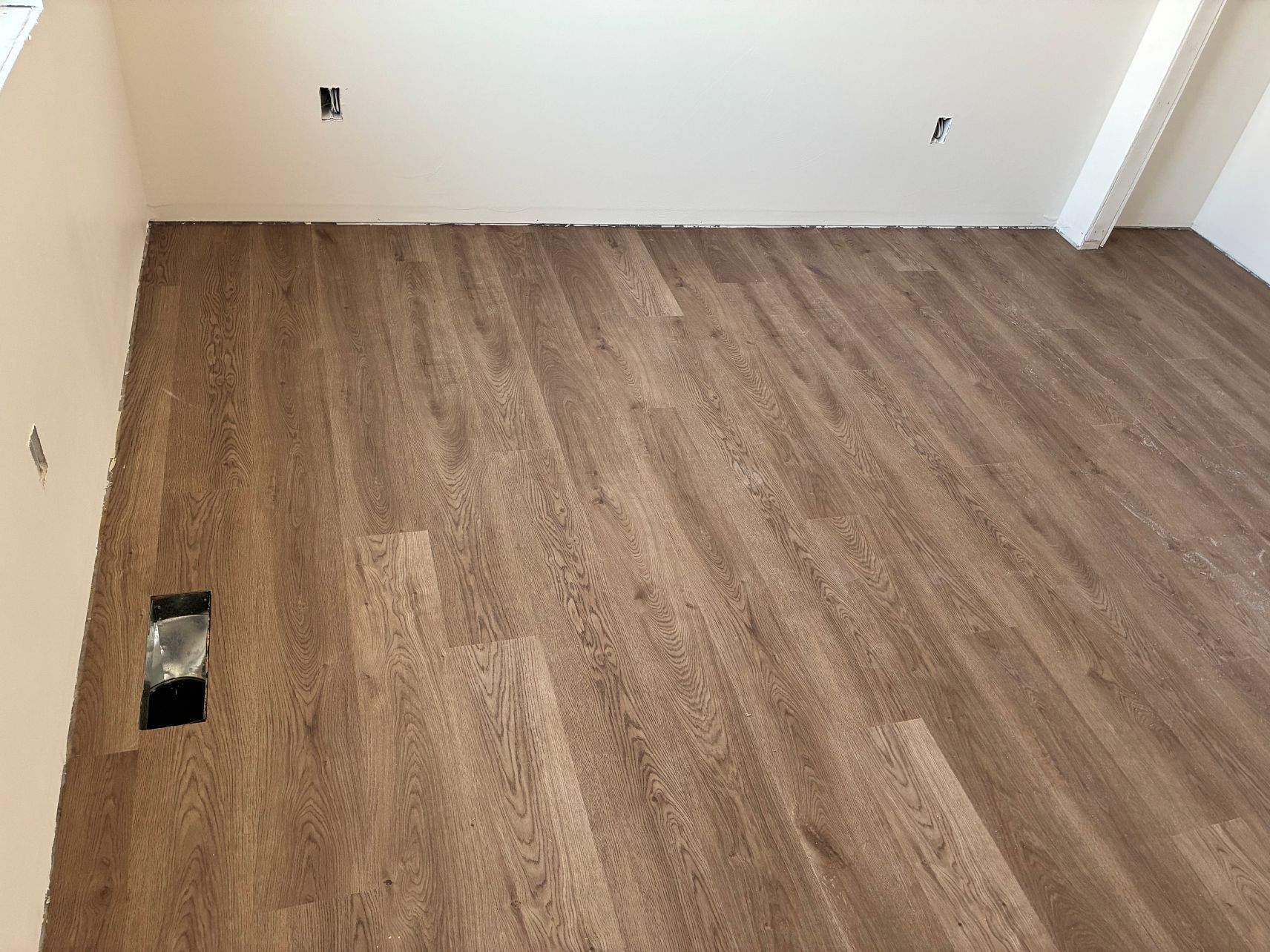Empty room with wood-look flooring and white walls, showing an electrical outlet opening near the floor.