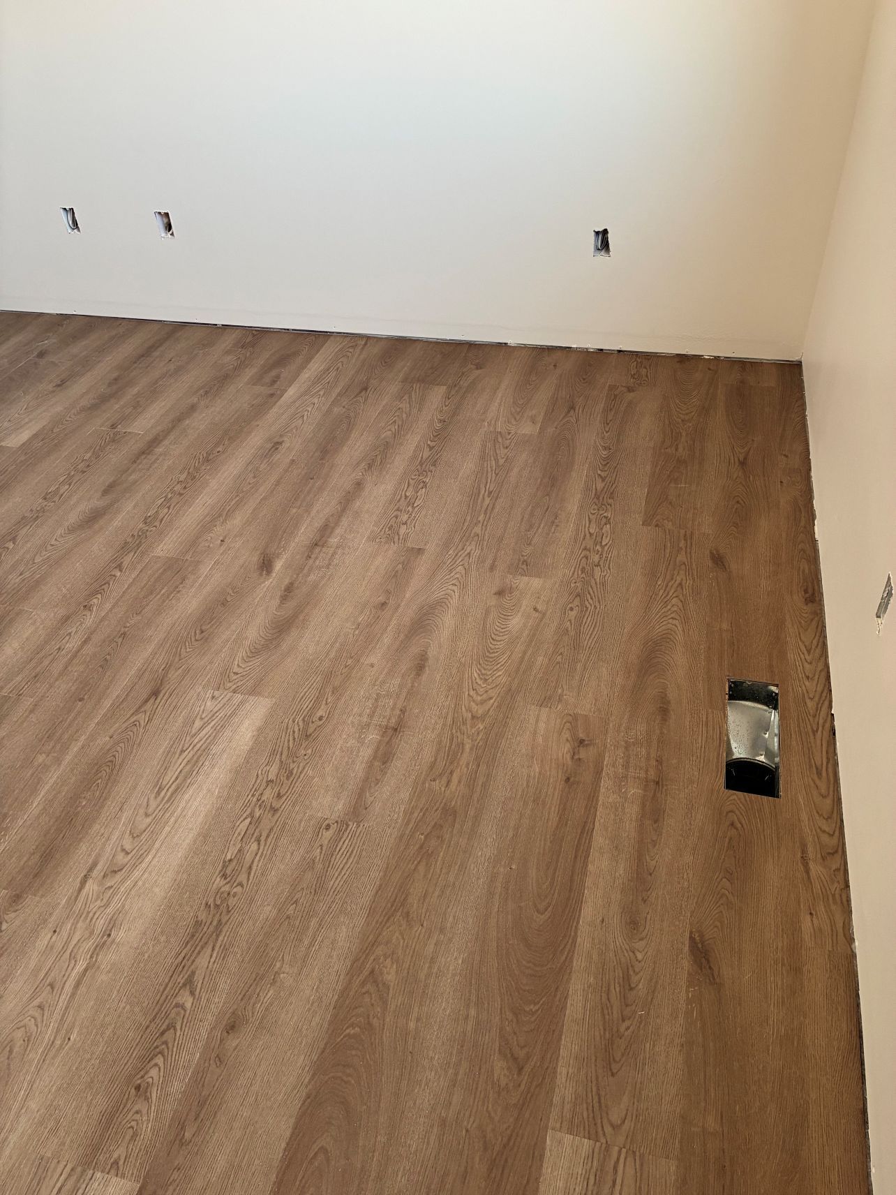 Empty room with wood flooring and white walls, showing electrical outlets and a floor cutout.