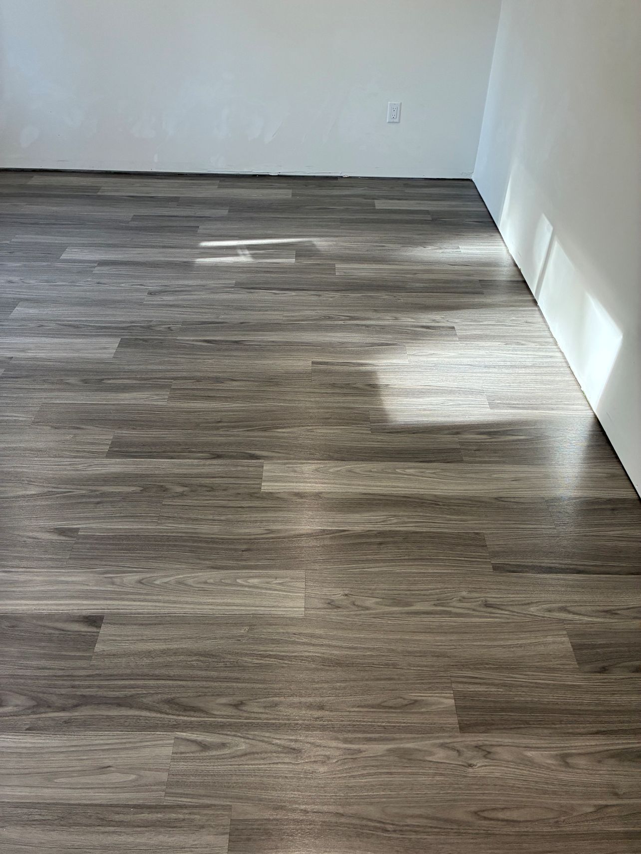 Empty room with gray wood-look flooring and white walls, lit by sunlight.