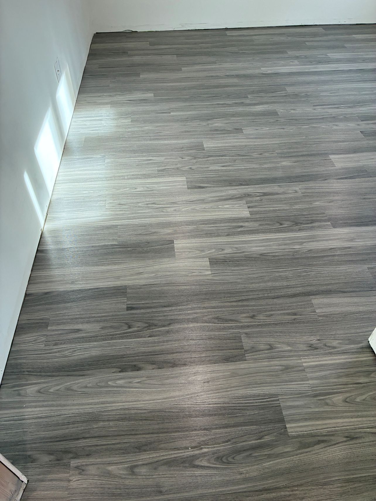 Empty room with gray wood-look flooring and white walls, lit by sunlight from the left.