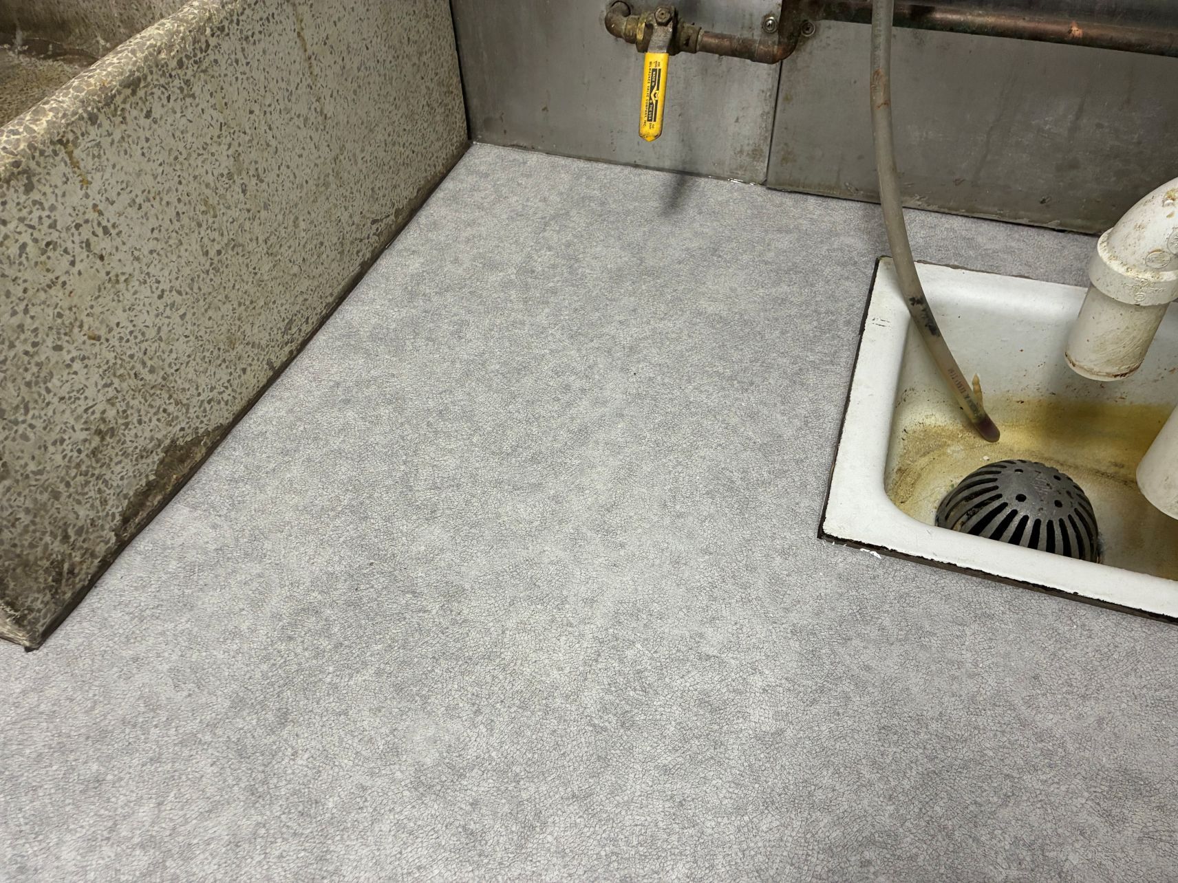 Gray utility floor with a drain and white sink pipes in a corner.