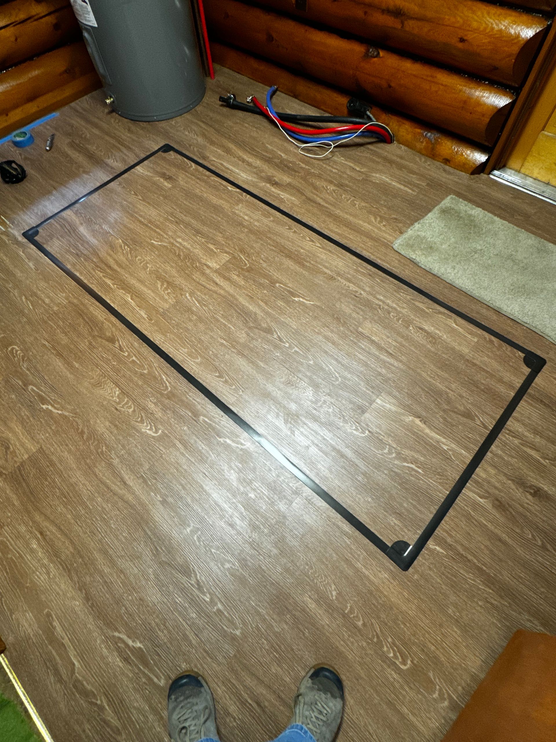 Wooden floor with a large rectangular outline of dark tape near a log wall and shoes at the bottom edge