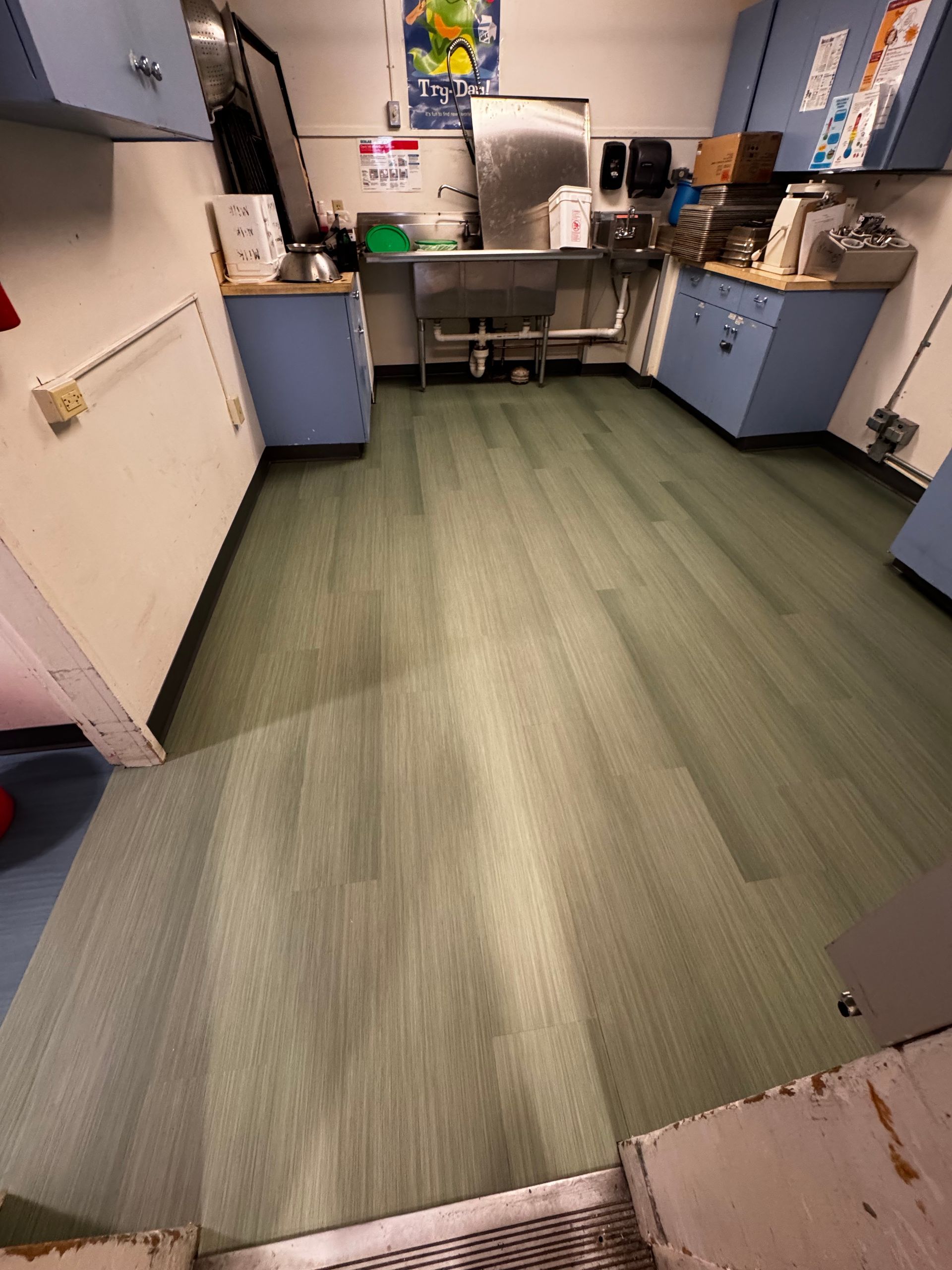 Empty kitchen with green floor, blue cabinets, and a stainless steel sink under bright lights
