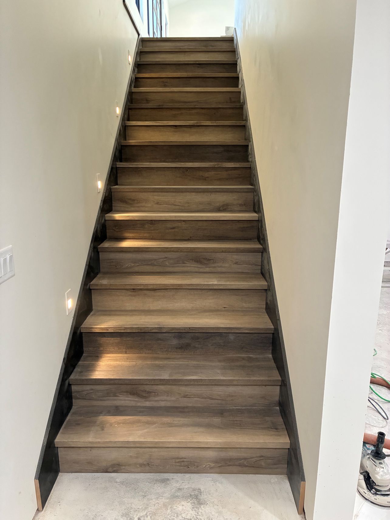Narrow wooden staircase with dark steps between white walls, viewed from the bottom.