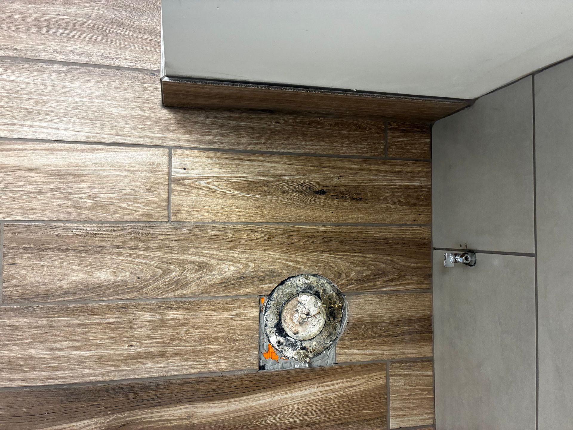 Circular floor drain set in wood-patterned flooring beside gray tile and a wall corner.