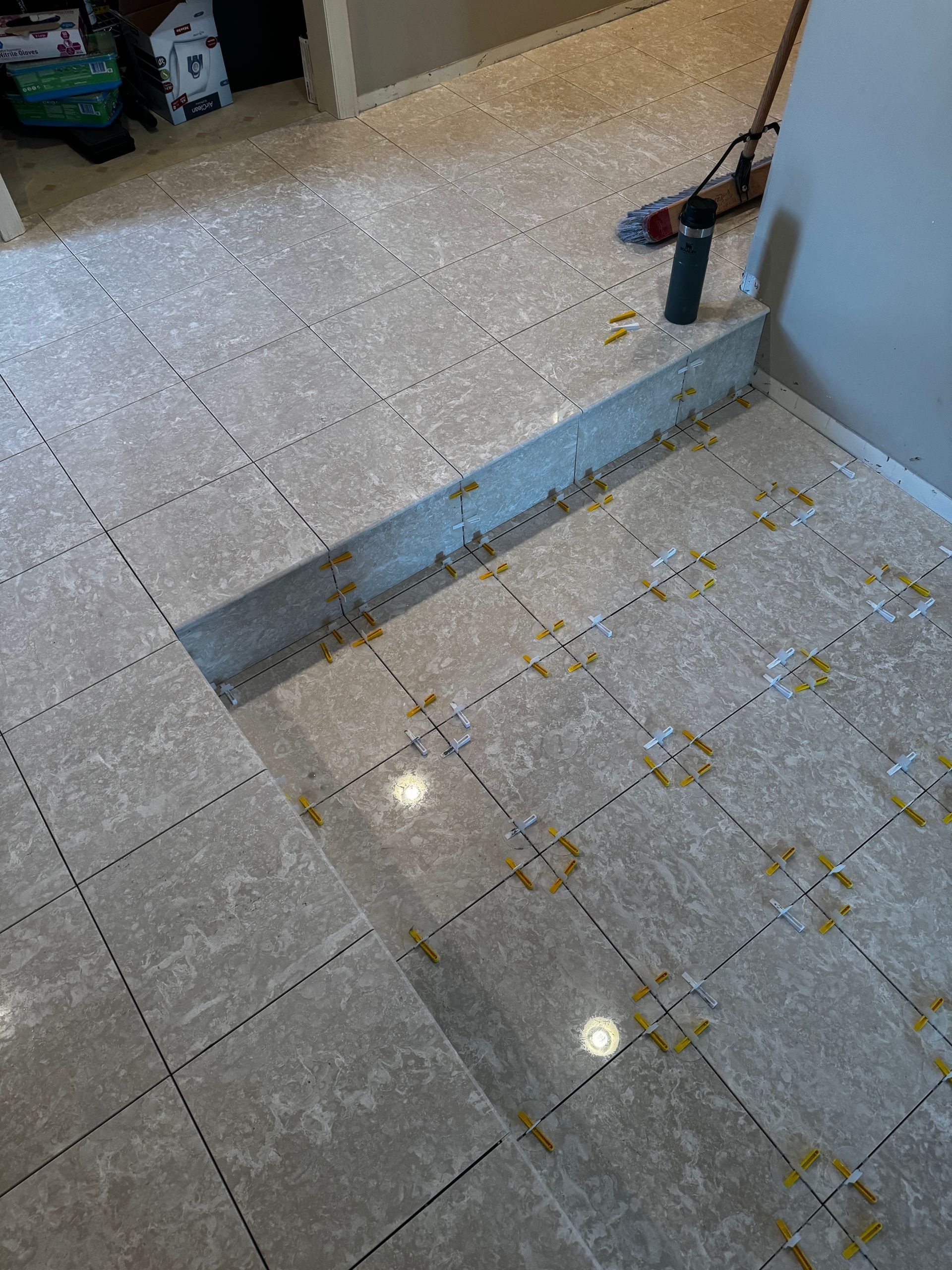 Indoor tiled floor with a recessed step or trench, scattered debris, and a tool leaning against the wall.