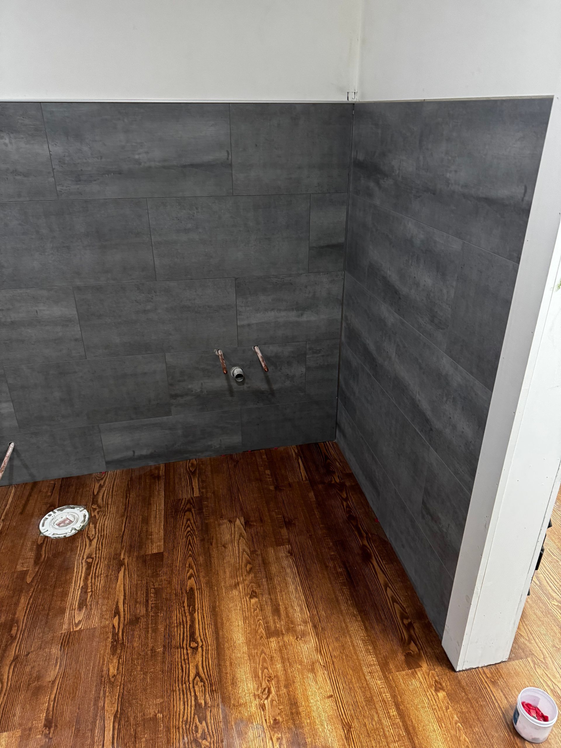 Empty tiled shower corner with gray walls and wood-look floor, showing a drain and water fixtures.