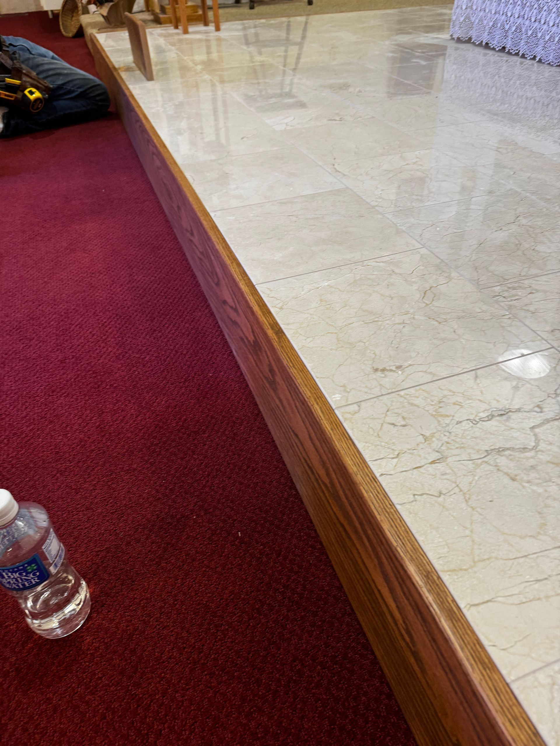 Church pew with red carpet, wood trim, and a water bottle on the floor beside a tiled aisle.