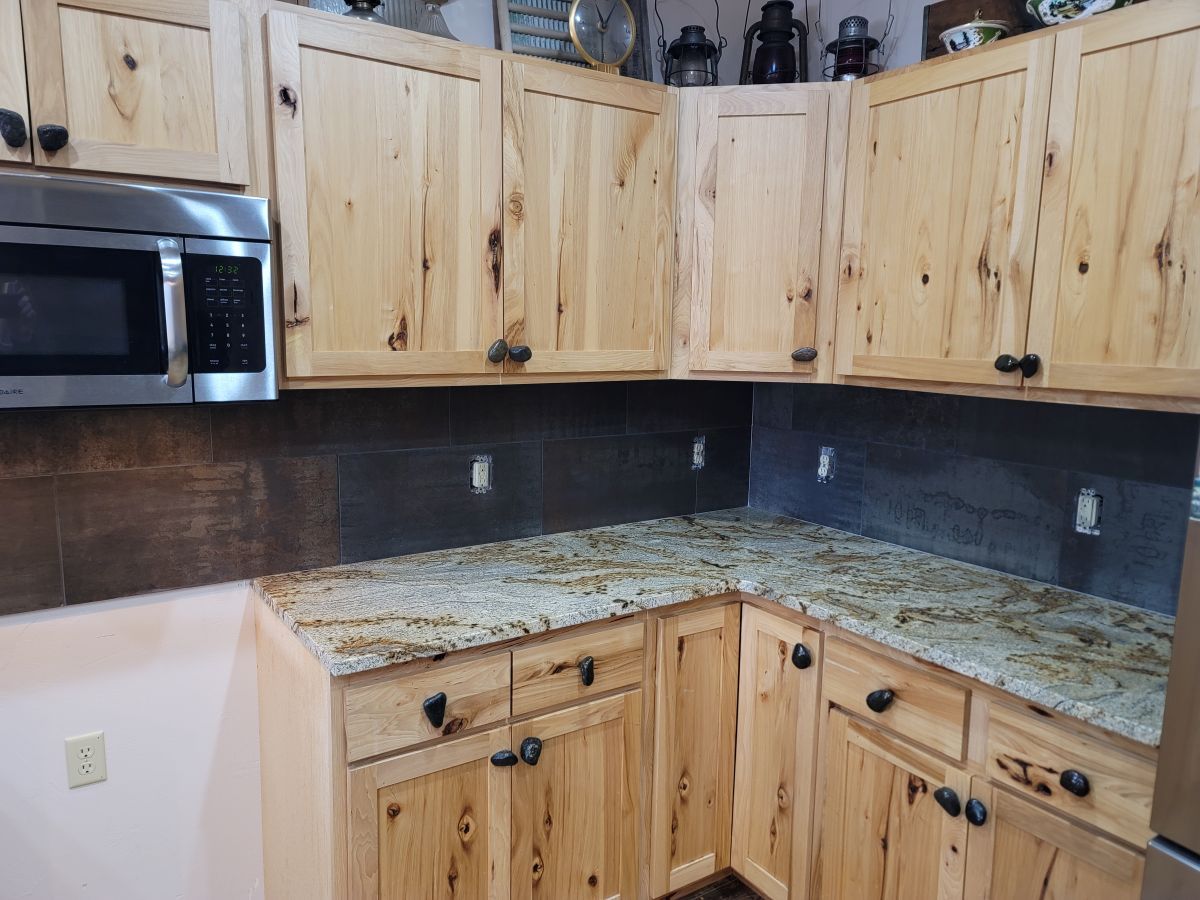 Kitchen corner with light wood cabinets, granite countertops, and a stainless microwave, lit by under-cabinet lighting