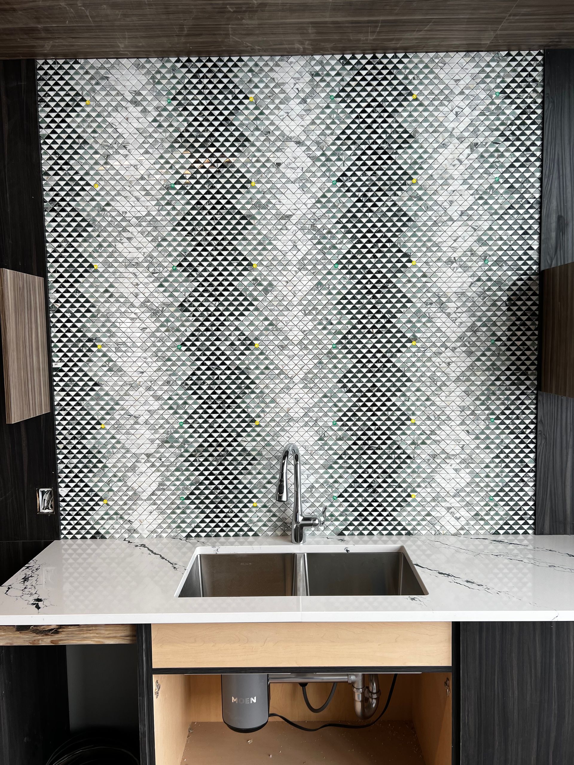 Kitchen sink beneath a metallic mosaic backsplash with speckled countertop and wooden cabinets below