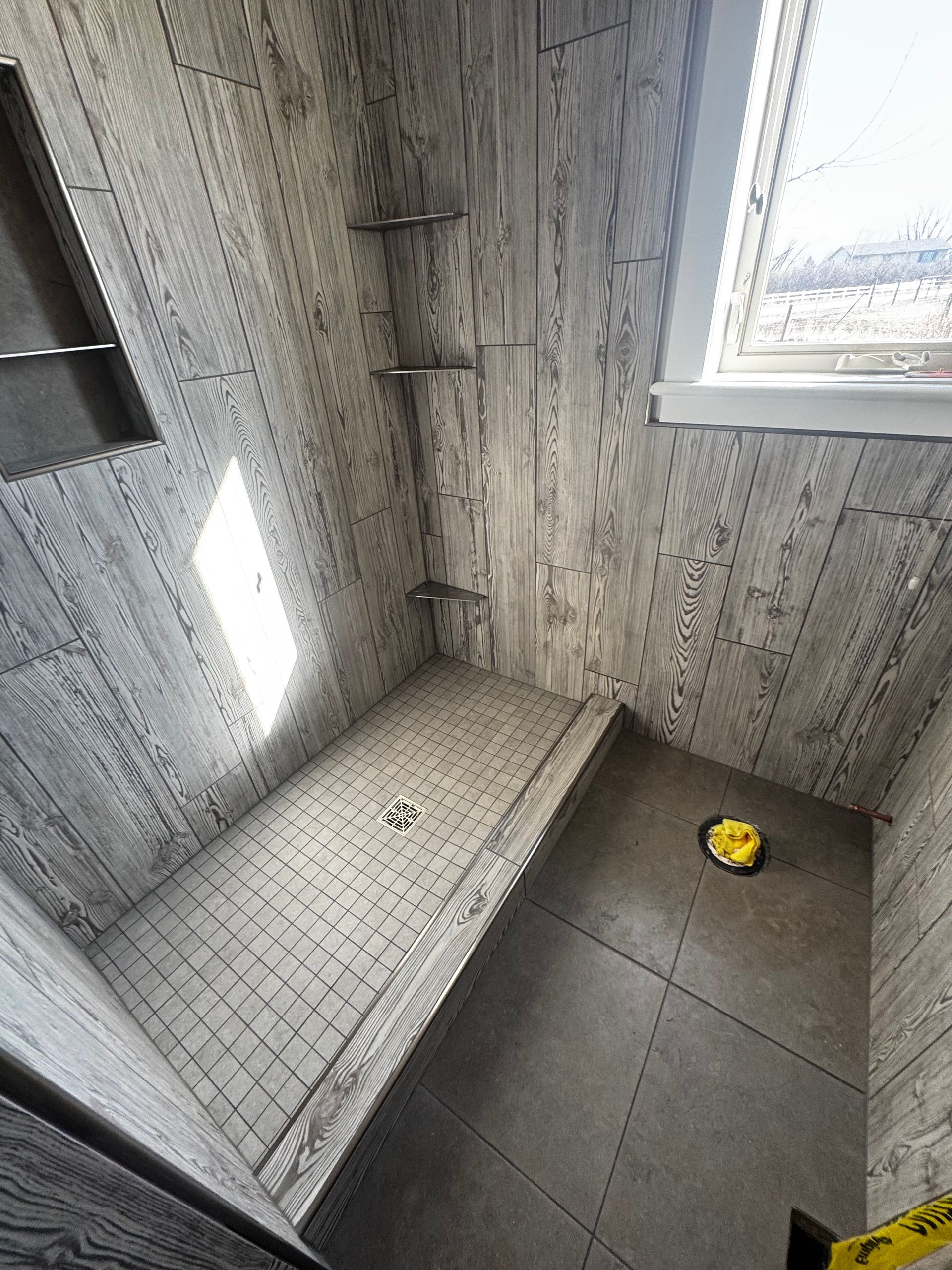 Small tiled shower with gray walls, dark floor, window, and a yellow bath toy in the corner.