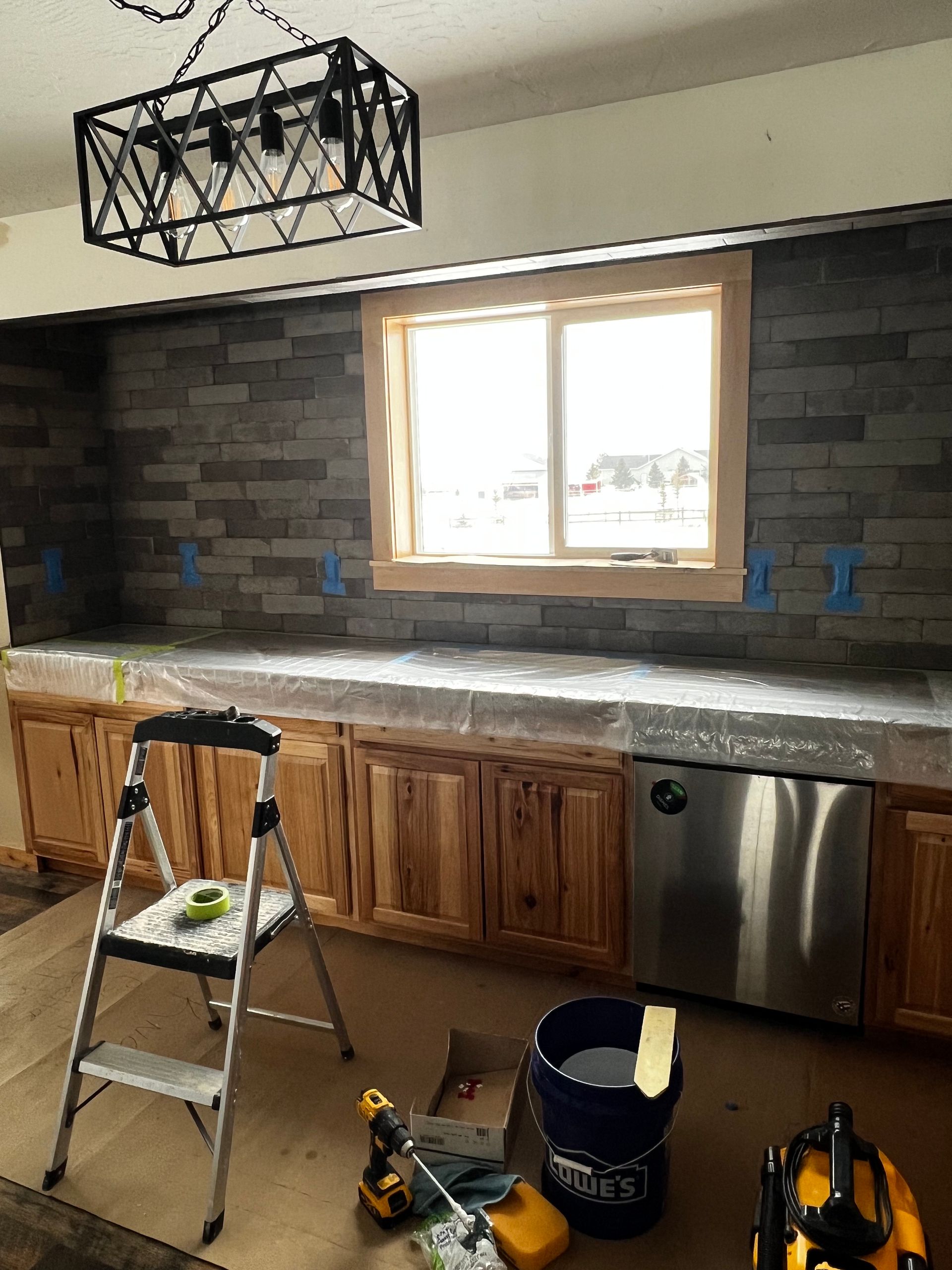 Kitchen under renovation with stone backsplash, wood cabinets, window, and tools on the floor.
