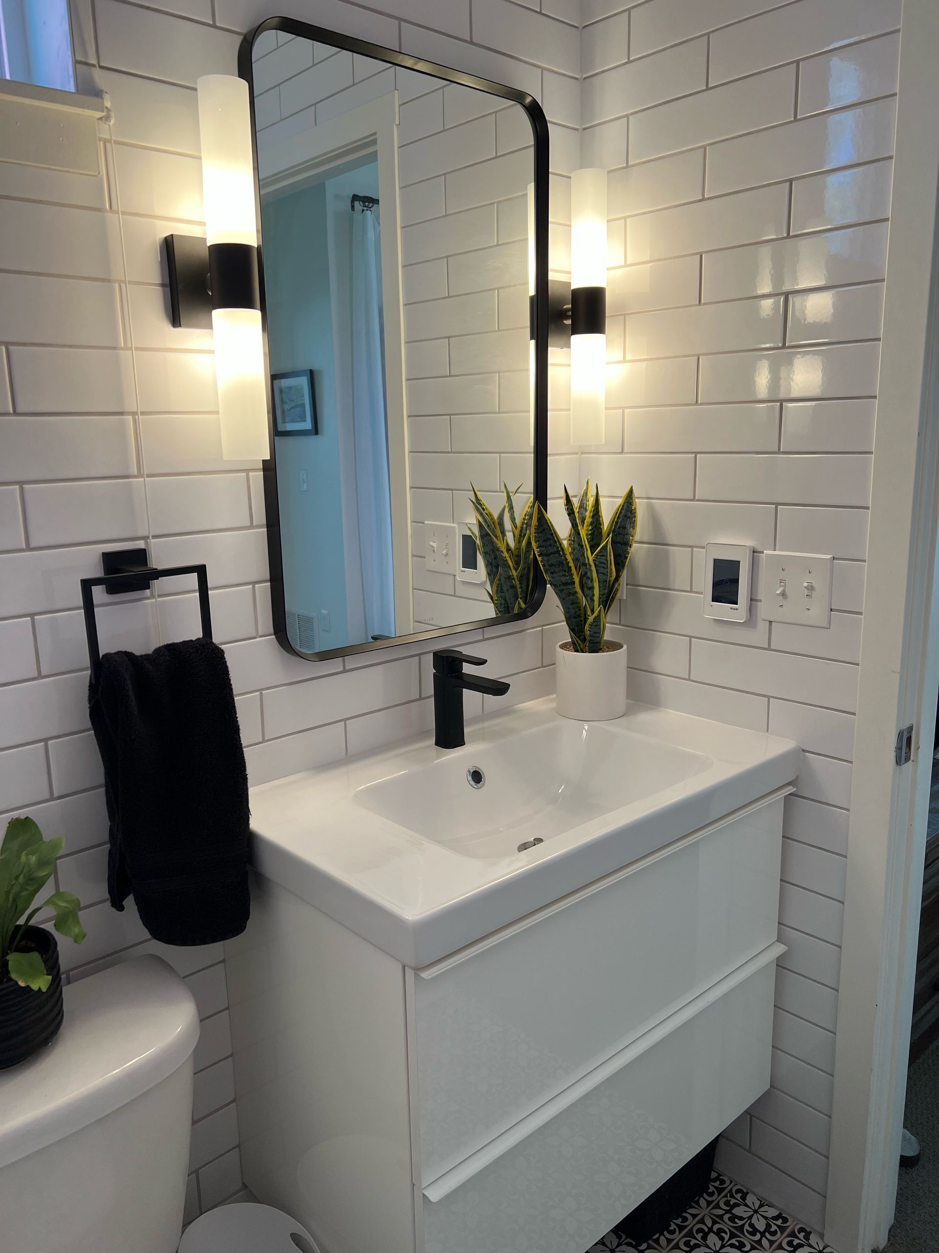 Small bathroom vanity with mirror, black fixtures, white tile, wall sconces, and a vase of yellow flowers.