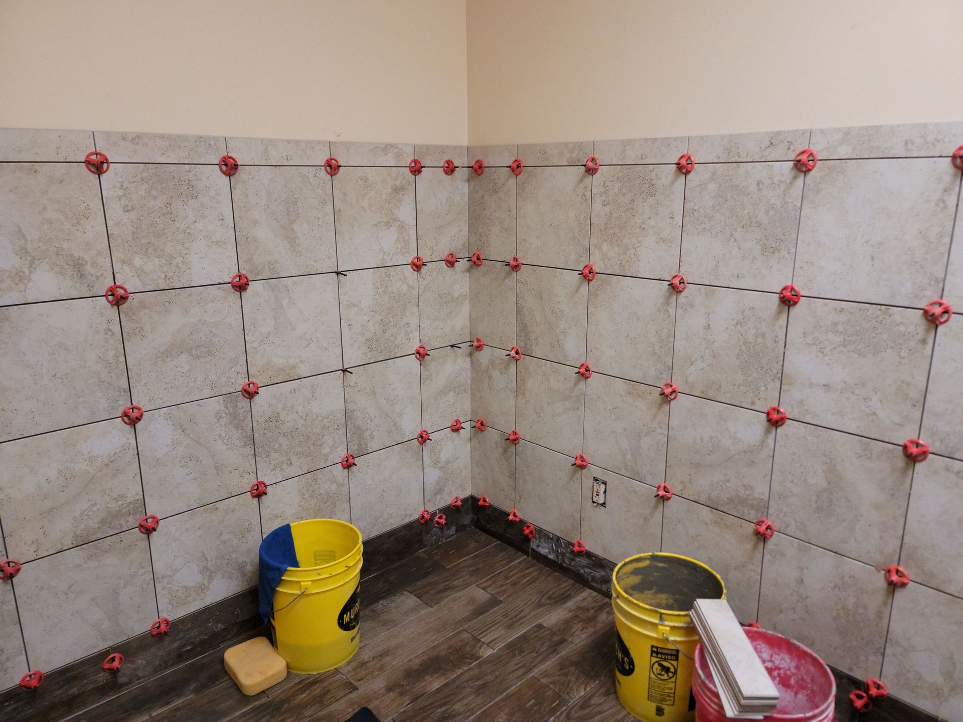 Tiled bathroom wall with red spacers, unfinished floor, and construction buckets and tools in the corner