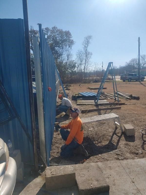 A man is kneeling down in front of a blue fence.