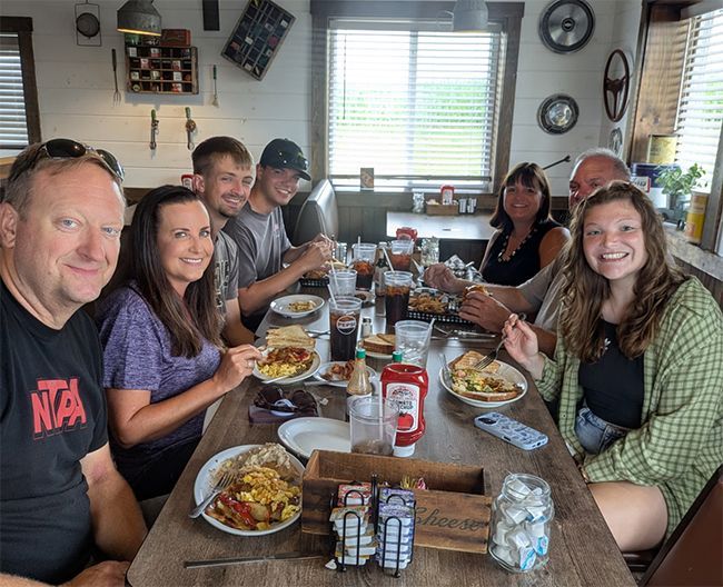 People gathered around a dining table with plates of food and drinks in a bright kitchen-dining room.