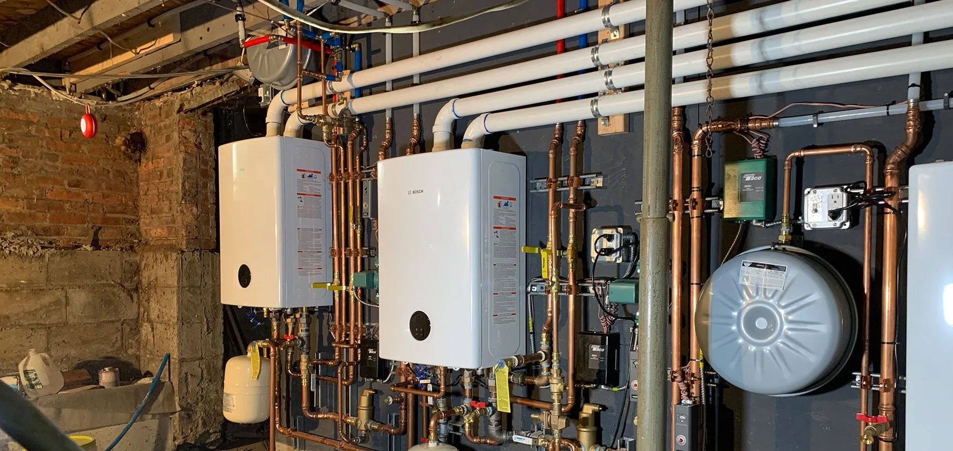 Piping and boilers in a basement. Copper pipes run between white boxes and other equipment.