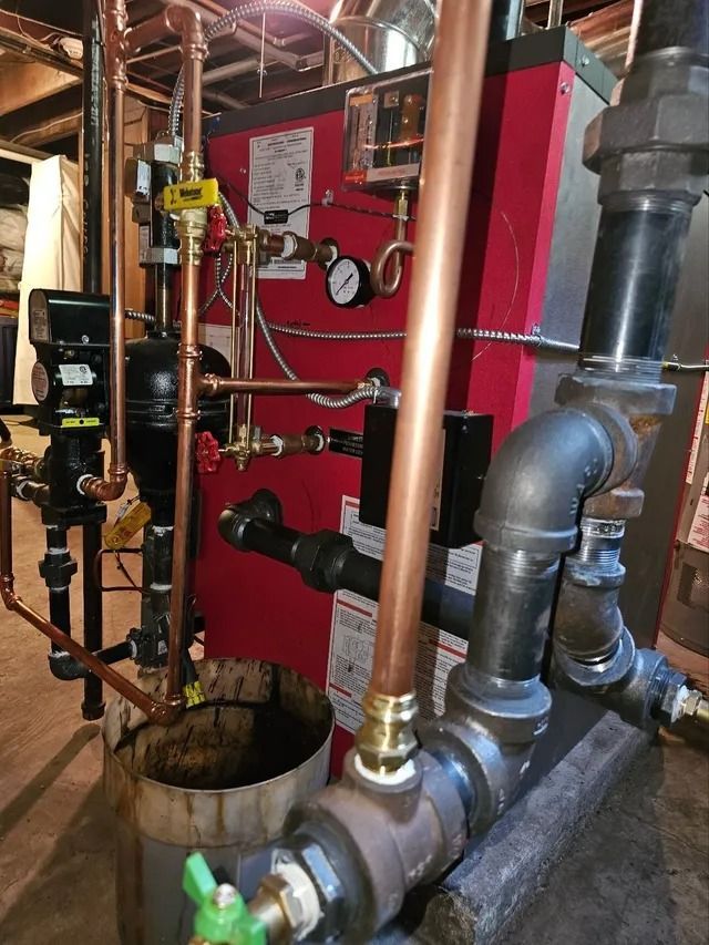 A red heating boiler with copper and black pipes in a basement setting.