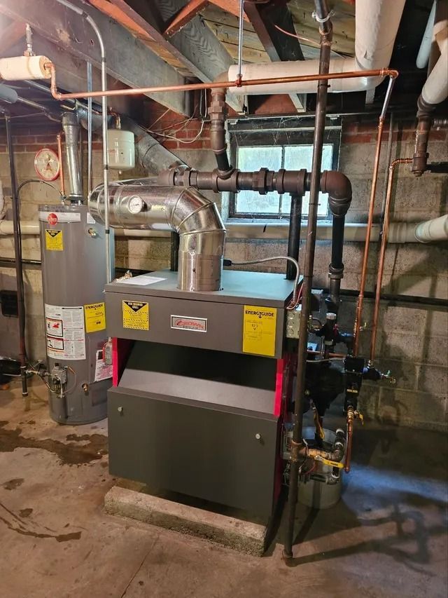 A furnace and water heater in a basement. Copper and black pipes are visible. Grey and red furnace. Concrete floor.