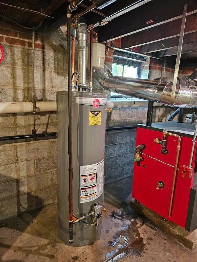Water heater and furnace in a basement. The water heater is gray. The furnace is red. Brick wall in the background.