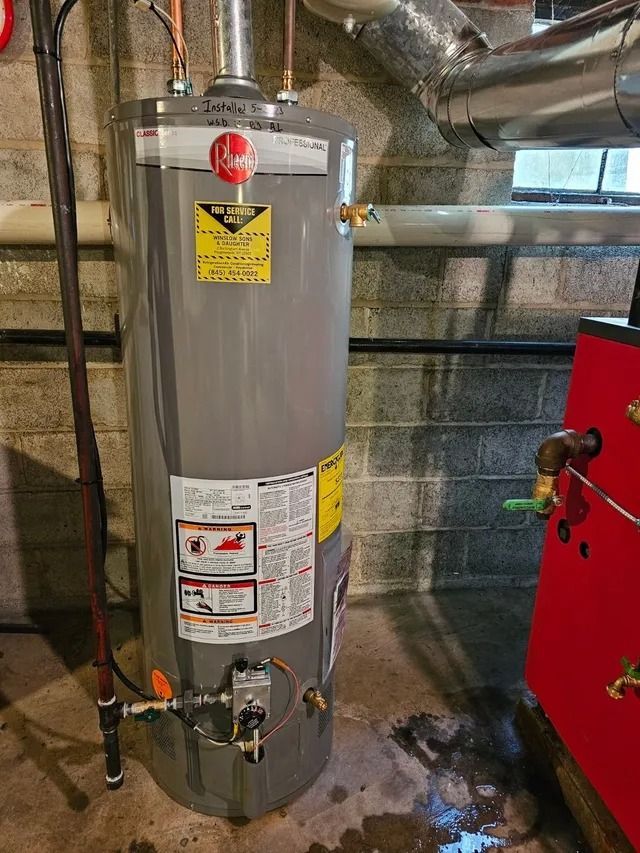 A tall, gray water heater in a basement setting with various pipes and a red furnace nearby.