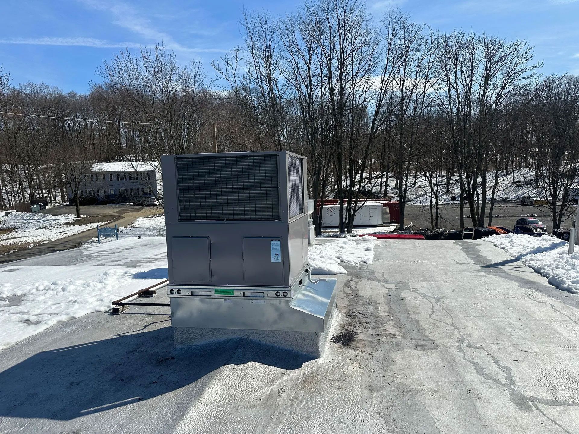 HVAC unit on a snowy rooftop with bare trees and a clear sky in the background.