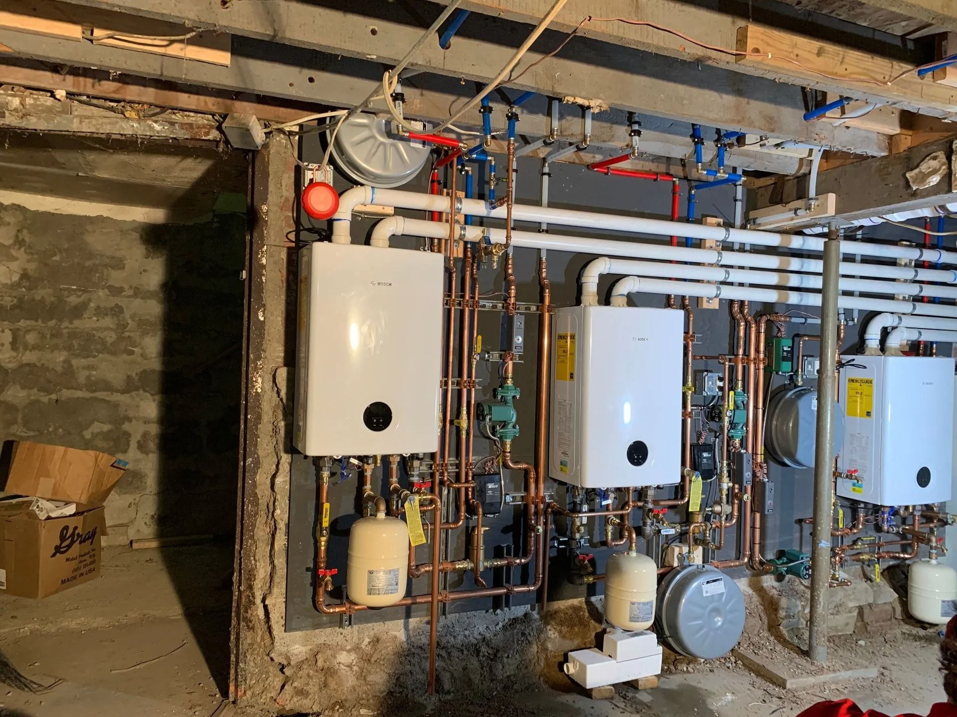 A basement with heating system components; white wall-mounted boilers, copper pipes, and a gray wall panel.
