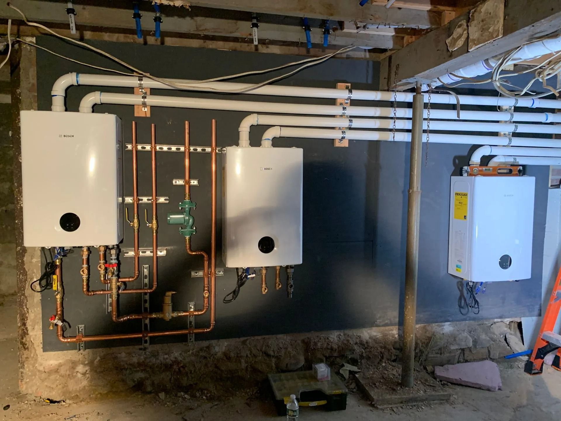 Three white wall-mounted boilers with copper pipes and white PVC pipes on a gray wall in a basement.