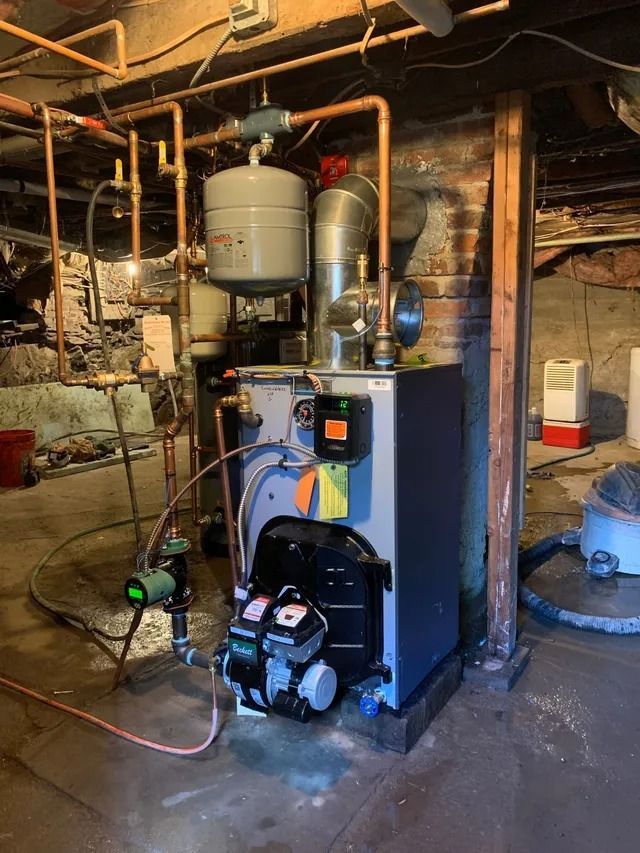 Furnace in a basement.  Dark blue furnace with silver pipes, tank, and a circular green device.  Exposed brick and wood.