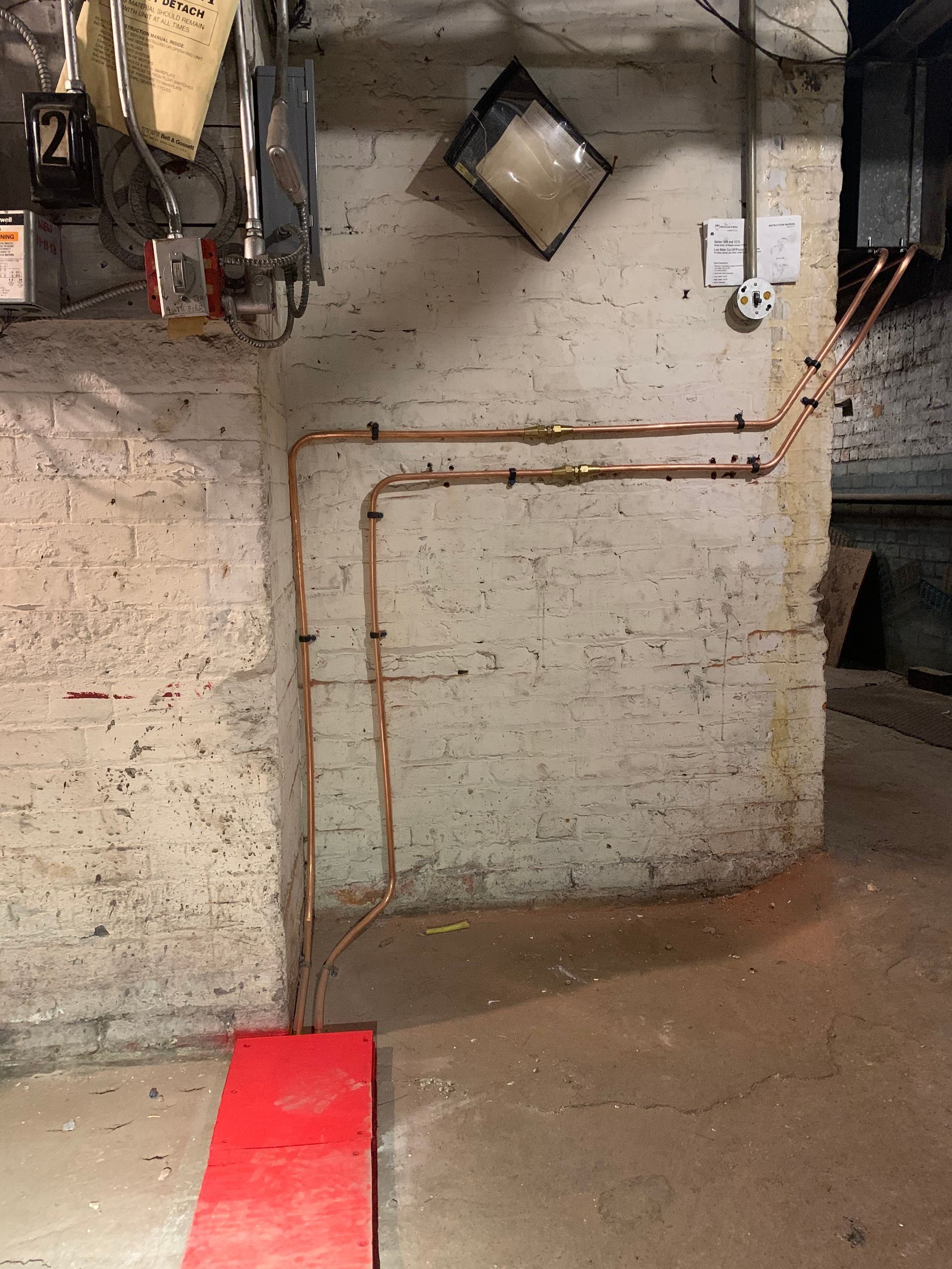 Copper pipes running along a white brick basement wall. A red object sits on the concrete floor.