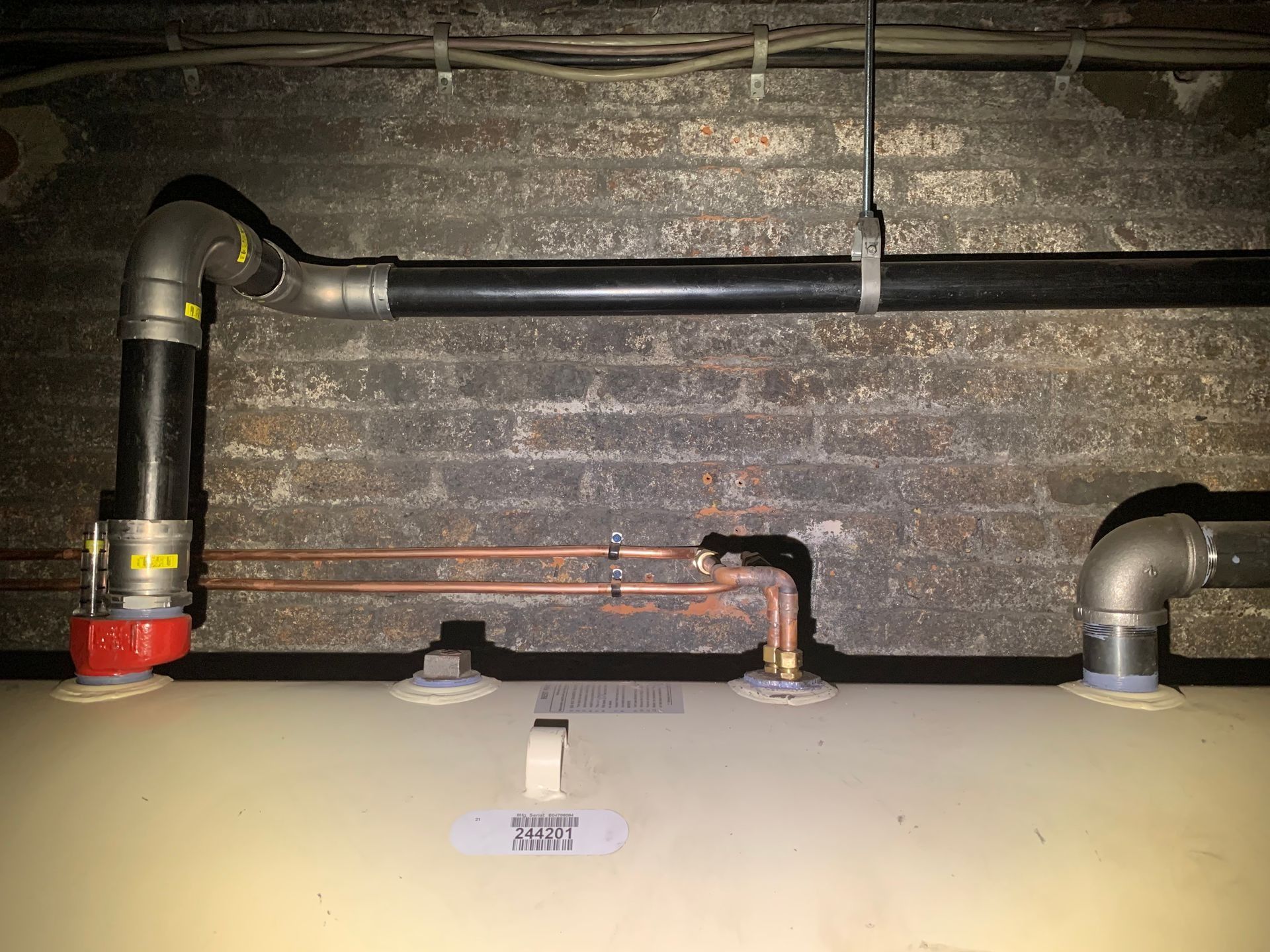 Pipes connected to a water heater in a basement with brick wall backdrop.