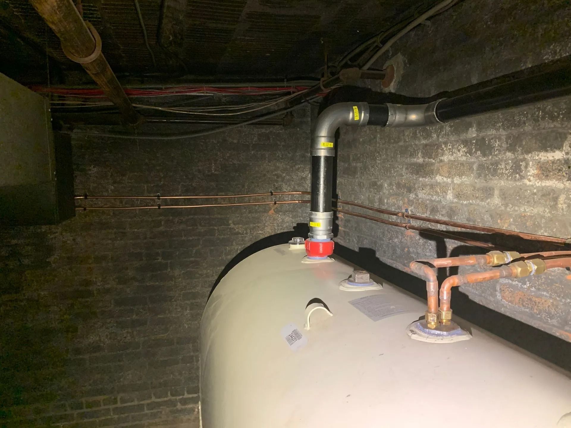 A cylindrical water tank with black pipes against a dark brick wall in a basement.