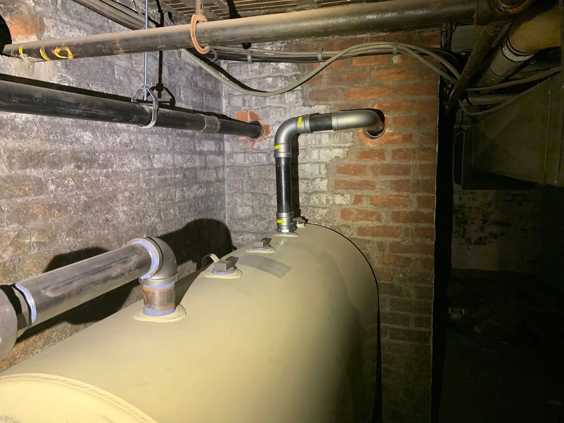 Oil tank with connected pipes in a basement against a brick wall.