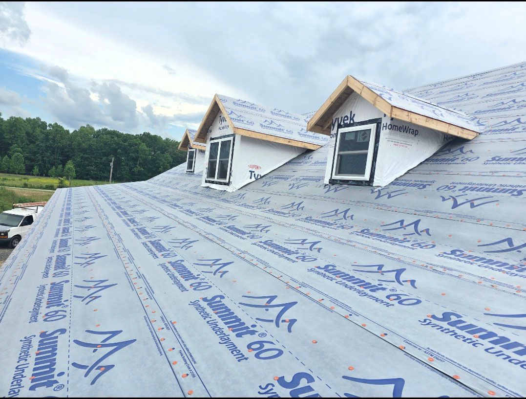 Roof under construction with blue underlayment and two dormer windows beneath cloudy skies