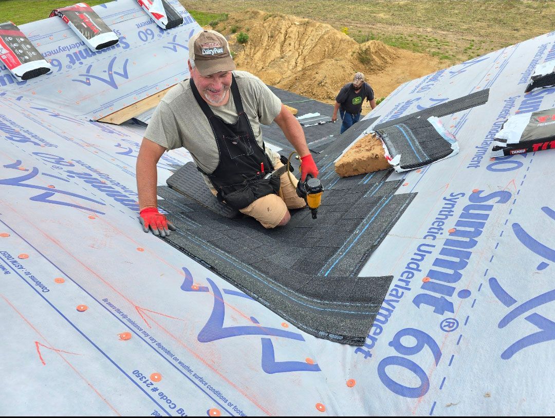 Worker kneeling on a roof, installing black underlayment amid gray roofing sheets and tools.