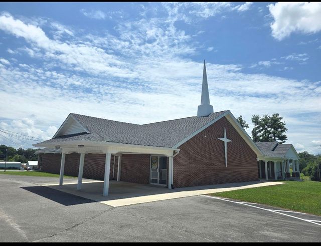 Small brick church with white cross and steeple under a blue sky, with a covered entrance and parking lot