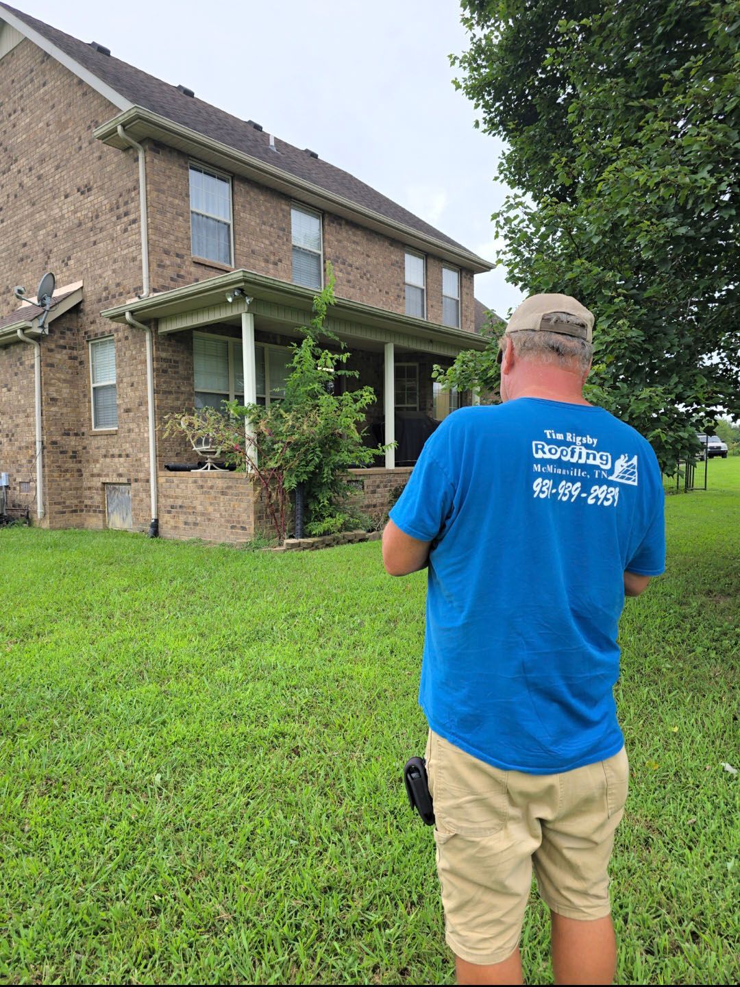 Man in blue shirt photographing a two-story brick house with a green lawn and porch