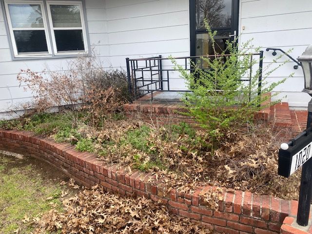 Brick garden bed with overgrown shrubs and fallen leaves beside a house porch and window.